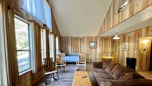 Un salon spacieux doté d’une grande fenêtre, de murs en bois, d’un canapé marron, d’une table basse en bois et d’un coin repas avec des chaises et une table.