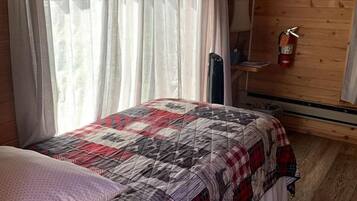 A wooden cabin room with a bed, curtains, a wall-mounted heater, and a framed picture.