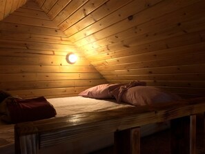 Un simple lit superposé en bois, avec un matelas et des oreillers, un petit sac et un luminaire au plafond.