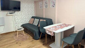 Ein gemütliches Zimmer mit einer Couch, einem kleinen Tisch mit einer blumigen Tischdecke, einem an der Wand montierten Fernseher und einer Wand mit geometrischem Muster.
