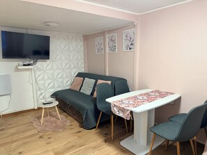 Ein gemütliches Zimmer mit einer Couch, einem kleinen Tisch mit einer blumigen Tischdecke, einem an der Wand montierten Fernseher und einer Wand mit geometrischem Muster.