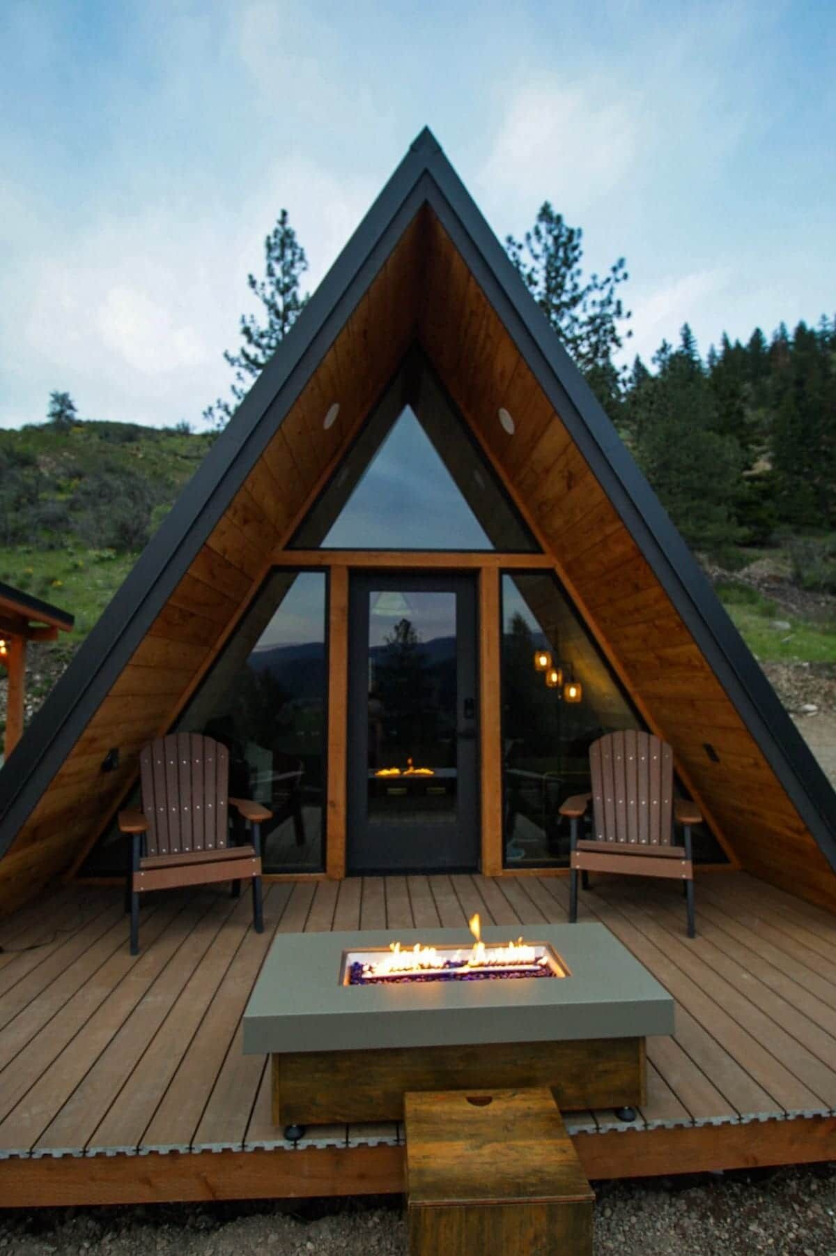 A triangular wooden cabin with a glass door, two Adirondack chairs, and a fire pit on the deck.