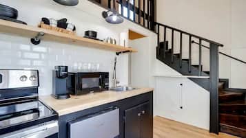Eine Küche mit Einbauofen, Mikrowelle, Kaffeemaschine und einem kleinen Kühlschrank. Über der Arbeitsplatte befindet sich ein Holzregal und rechts eine Treppe.