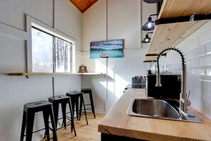 A kitchen with a wooden countertop, a stainless steel sink, black stools, a window with a view, and a painting on the wall.
