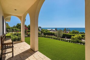 A covered patio with a view of the sea, a lawn, and a garden.
