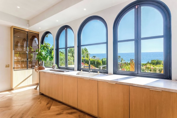 A kitchen with large windows, wooden cabinets, and a view of the sea.