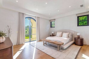 A bedroom with a large bed, a bench, a nightstand, a potted plant, and a view of greenery outside.