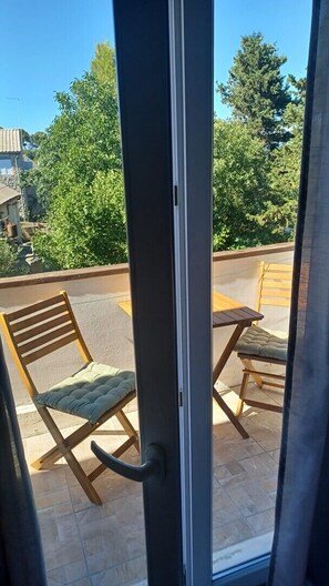 Ein Balkon mit zwei Holzstühlen und einem Tisch, einer Glastür und Blick auf Bäume und Gebäude.
