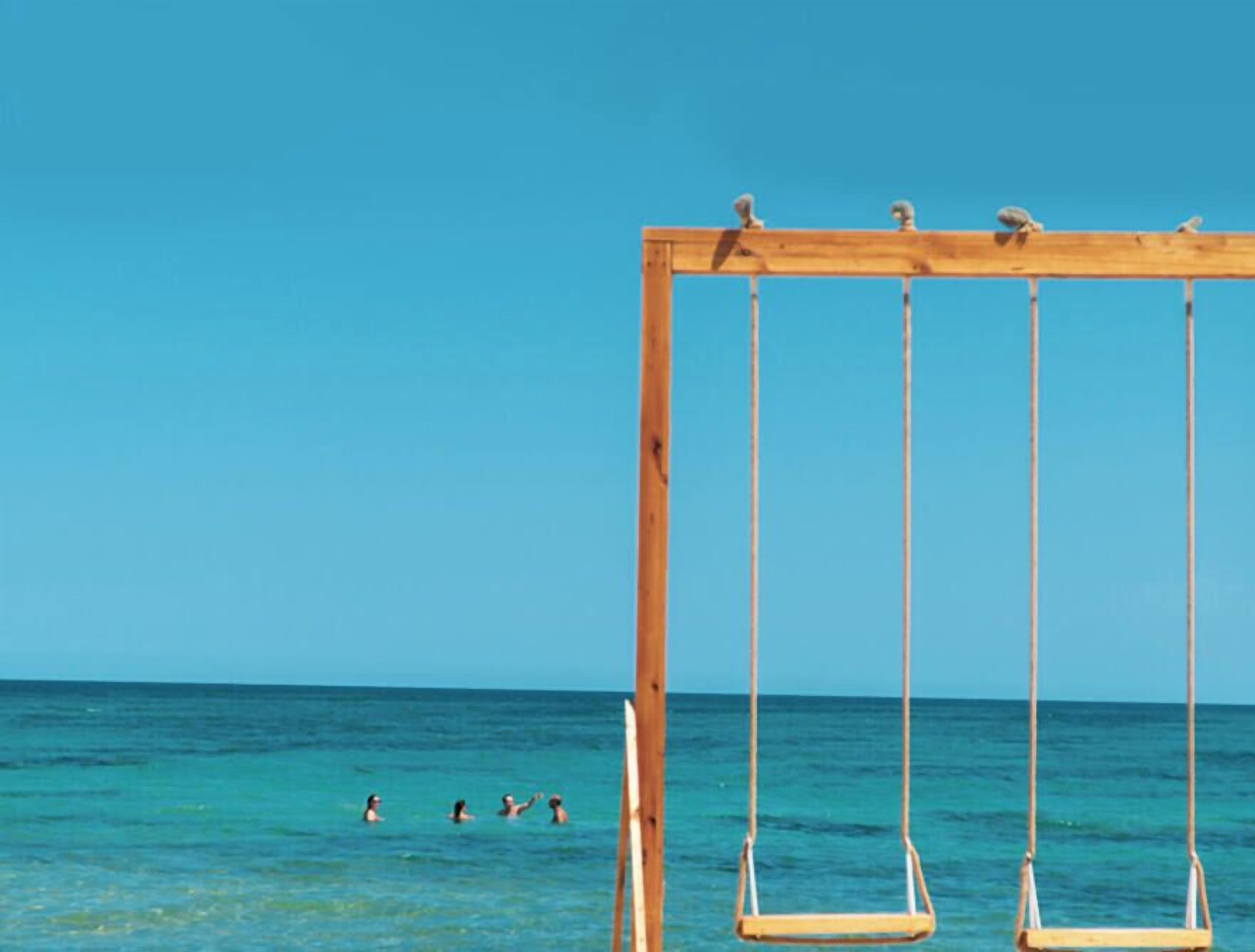 Une balançoire en bois installée au bord de la mer, avec une eau d’un bleu limpide et un ciel dégagé.