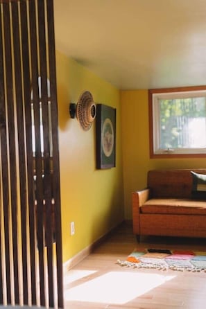 A room with a wooden floor, a sofa, a wall-mounted fan, and a window with a view of greenery. - The Catskill | Three Retreats One Creekside Escape (Jefferson Heights)