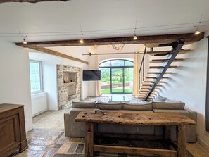 A spacious living area with a stone fireplace, a large window offering a view of greenery, and a wooden staircase. - La Louverie | Private Jacuzzi | View of the Gorges du Tarn (Mostuéjouls)