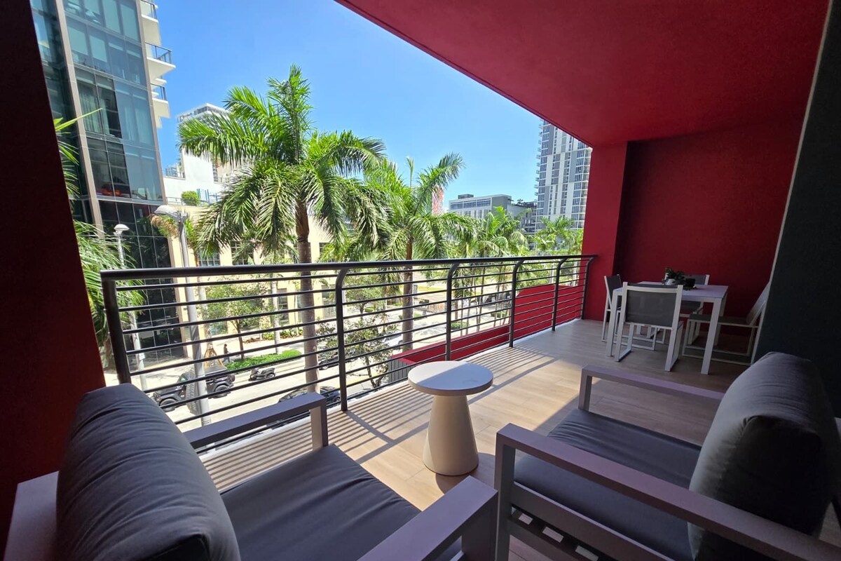 A balcony with a view of palm trees and buildings, a table, chairs, and a bench.