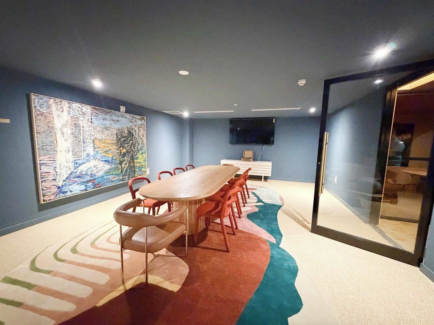 Un salón de conferencias con una larga mesa, sillas, un gran cuadro abstracto, un televisor y una alfombra con un motivo de olas.