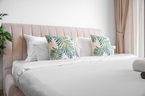 Ein Bett mit weißer Bettwäsche und zwei dekorativen Kissen mit Blätermuster, ein beige Kopfschild und eine Pflanze auf der linken Seite.