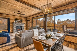 L’intérieur chaleureux d’un chalet avec une cheminée en pierre, un coin repas aménagé pour quatre personnes et une grande fenêtre donnant sur les arbres aux couleurs automnales.