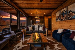 A cozy cabin interior with a wooden ceiling, a large window overlooking a scenic view, a dining table with candles, and a blue sofa. - Panoramic Views Cabin/ HotTub/ Arcade/ Ski/ Pickleball/ Golf in Gatlinburg,TN (Gatlinburg)