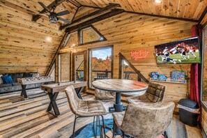 Ein gemütliches Zimmer mit Holzbalkendecke, einem Kicker, einem runden Tisch mit Stühlen und einem Fernseher, der ein Fußballspiel zeigt.