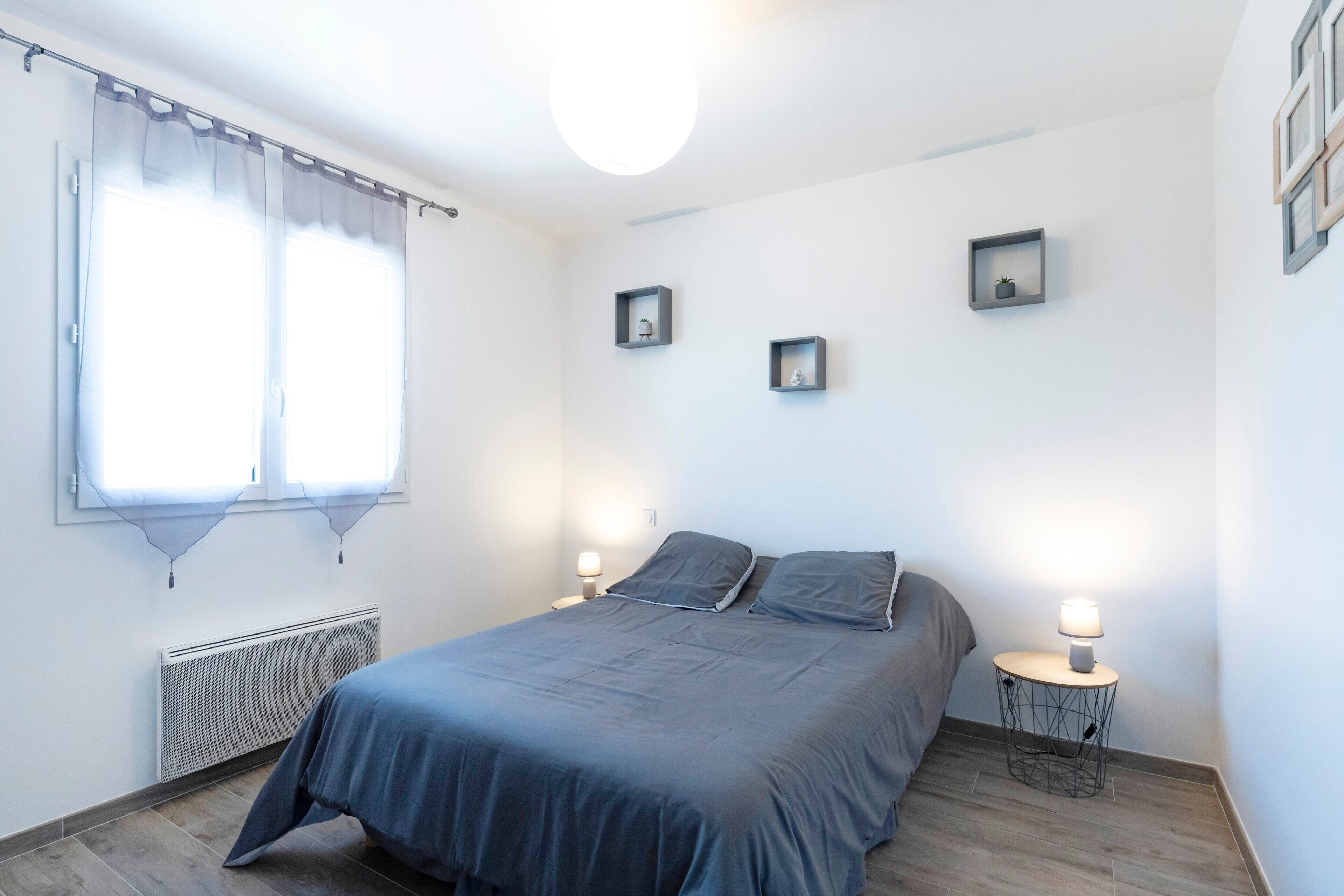 Un dormitorio con cama, dos mesitas de noche con lámparas, un radiador y una ventana con cortinas.