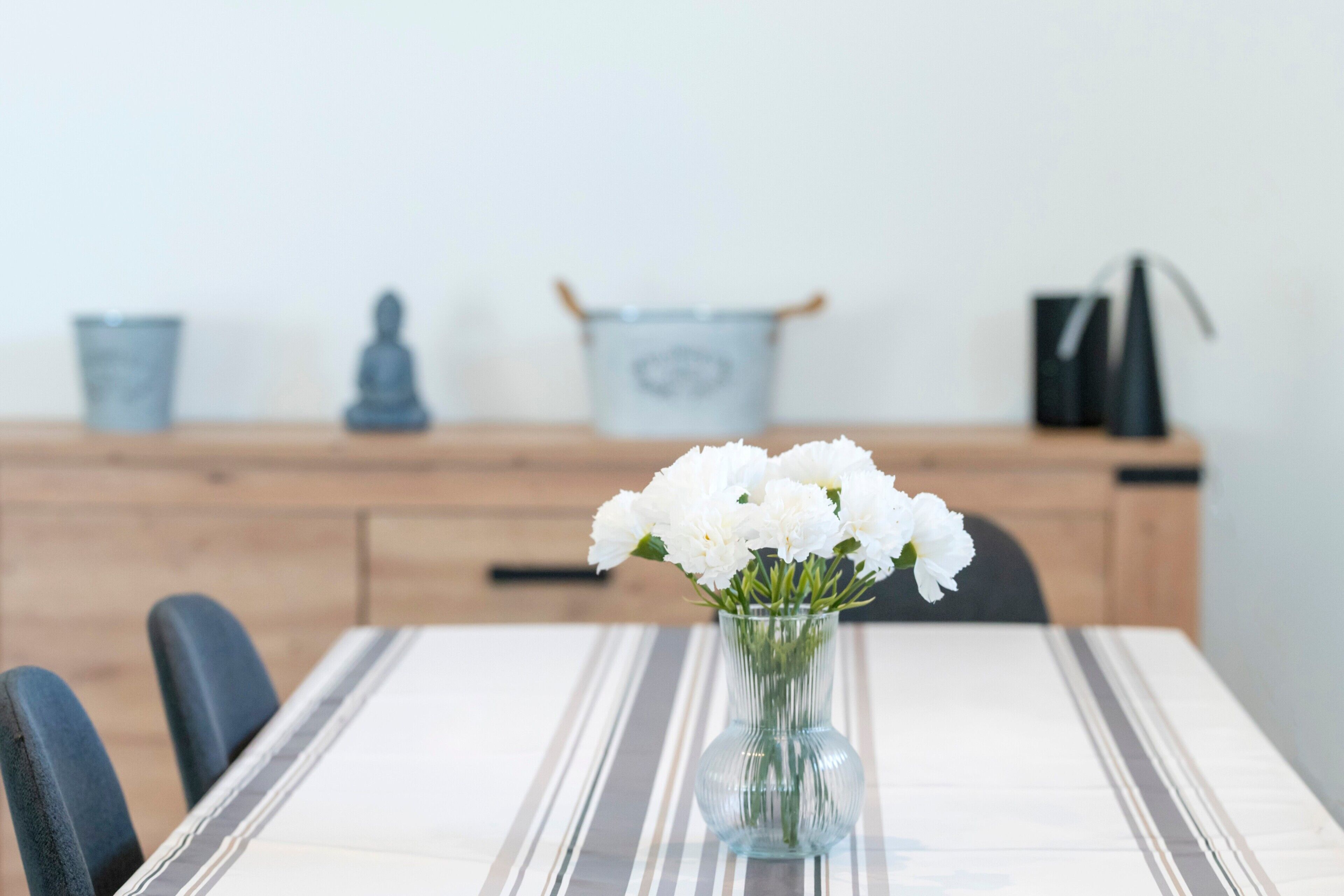 Una mesa de comedor con un jarrón de flores blancas, un mantel a rayas y un armario de madera al fondo.