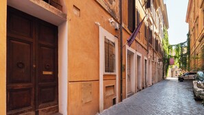 A narrow cobblestone street with traditional buildings, a parked car, and a visible door with a window. - Via del Corso Quiet Design Studio | Elevator (Roma)