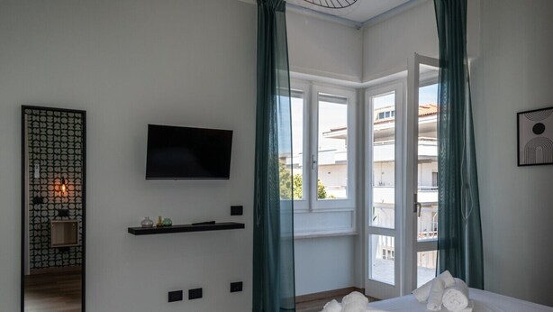 A hotel room with a bed, a TV, a window with curtains, a wall-mounted shelf, and a decorative mirror. - Allegra House - Three-Bedroom Apartment (Alba Adriatica)
