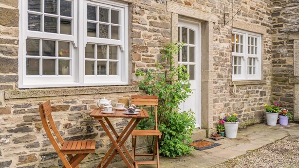 Un bâtiment en pierre avec deux fenêtres à cadre blanc, une petite table avec un service à thé et des plantes en pot.