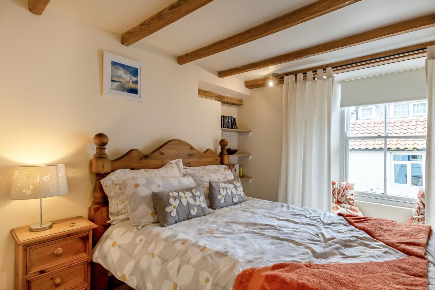 A bedroom with a wooden headboard, a bedside table with a lamp, a bed with floral bedding, a window with curtains, and a view of buildings outside.
