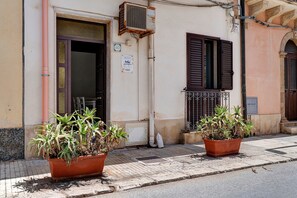 Un edificio con una porta, una finestra con persiane e un cartello. Due piante in vaso sono posizionate davanti all'edificio.