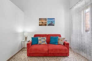 Ein rotes Sofa mit gemusterten Kissen, ein kleiner Tisch mit einer Lampe und zwei gerahmte Bilder an der Wand.
