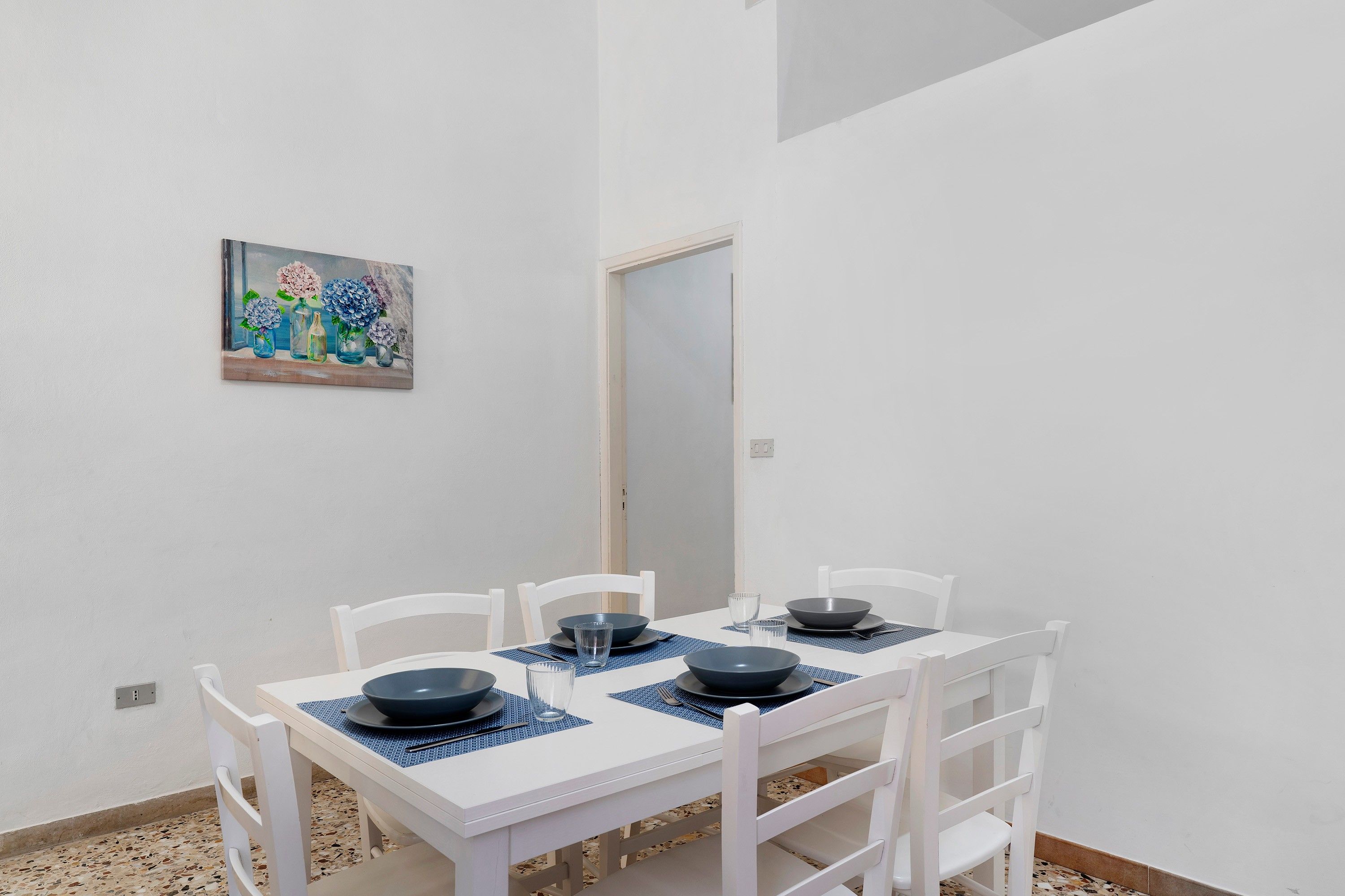 Un coin repas avec une table et des chaises blanches, un tableau au mur et une porte à l’arrière-plan.
