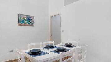 Un coin repas avec une table et des chaises blanches, un tableau au mur et une porte à l’arrière-plan.