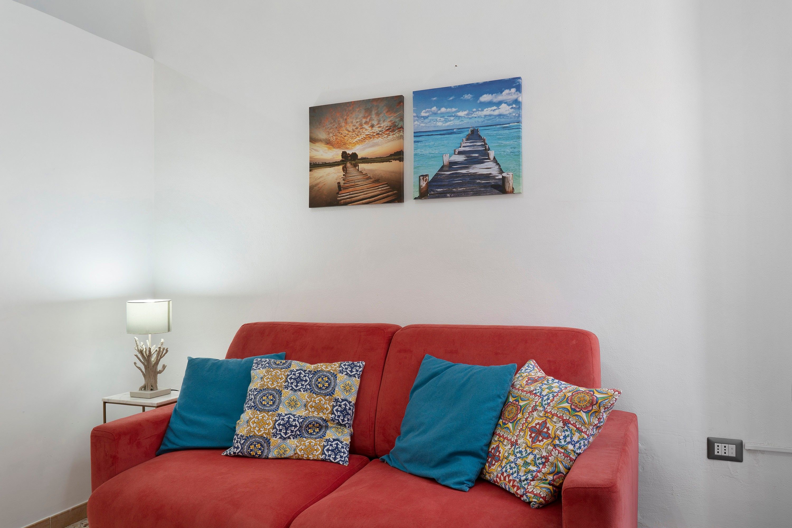 Ein rotes Sofa mit gemusterten Kissen, ein kleiner Tisch mit einer Lampe sowie zwei an der Wand hängende Bilder, die einen Sonnenuntergang und einen Steg zeigen.