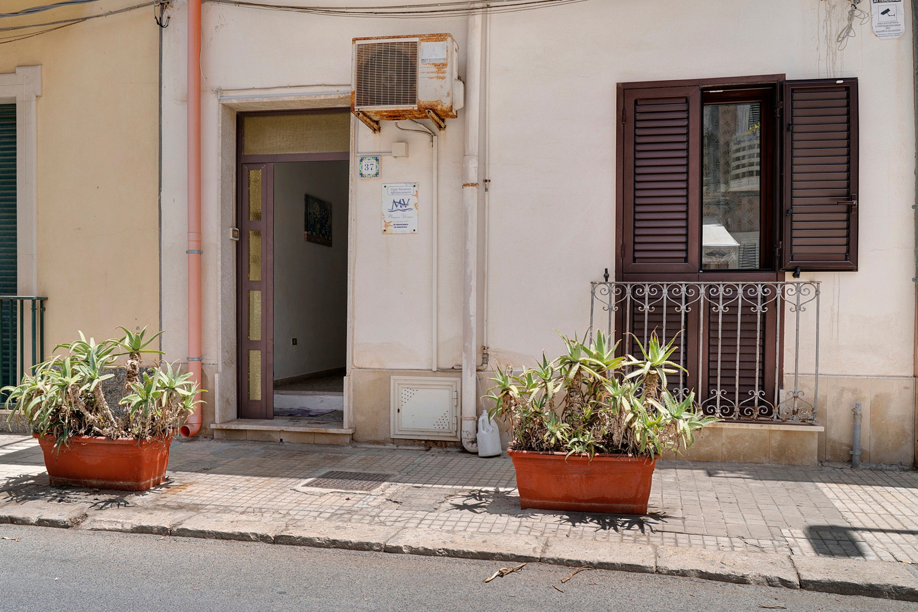 Un bâtiment à la façade beige, avec une porte marron et une fenêtre dotée de volets marrons. Deux plantes en pot sont placées devant le bâtiment.
