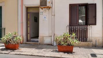Un bâtiment à la façade beige, avec une porte marron et une fenêtre dotée de volets marrons. Deux plantes en pot sont placées devant le bâtiment.