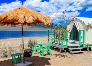 A beachside tent with a straw umbrella, green chairs, and a small table.