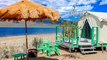 A beachside tent with a straw umbrella, green chairs, and a small table.