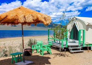 Une tente installée sur la plage, avec un parasol en paille, des chaises vertes et une petite table.
