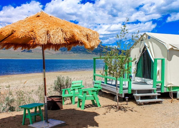 A beachside tent with a straw umbrella, green chairs, and a small table.
