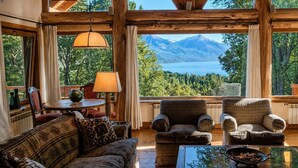 Un interior acogedor con un amplio ventanal con vistas a un lago y montañas, vigas de madera, un sofá, sillones y una mesa con un cuenco de nueces.