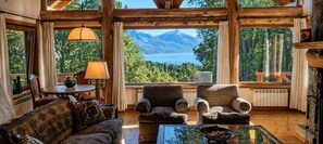 A cozy interior with a large window overlooking a lake and mountains, wooden beams, a sofa, armchairs, and a table with a bowl of nuts.