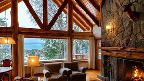 Interior acogedor de una cabaña con chimenea de piedra, vigas de madera y grandes ventanas que ofrecen vistas al exterior.