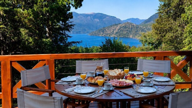 Una terraza de madera con una mesa redonda dispuesta para el desayuno, con vistas a un lago y montañas.