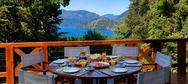 A wooden deck with a round table set for breakfast, overlooking a lake and mountains.
