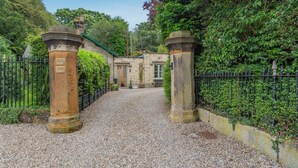 A gravel driveway leading to a stone building with a green roof, surrounded by a black metal fence and lush greenery. - 1 Bed in Great Ayton (oc-h44914) (Great Ayton)