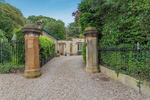 A gravel driveway leading to a stone building with a green roof, surrounded by a black metal fence and lush greenery. - 1 Bed in Great Ayton (oc-h44914) (Great Ayton)