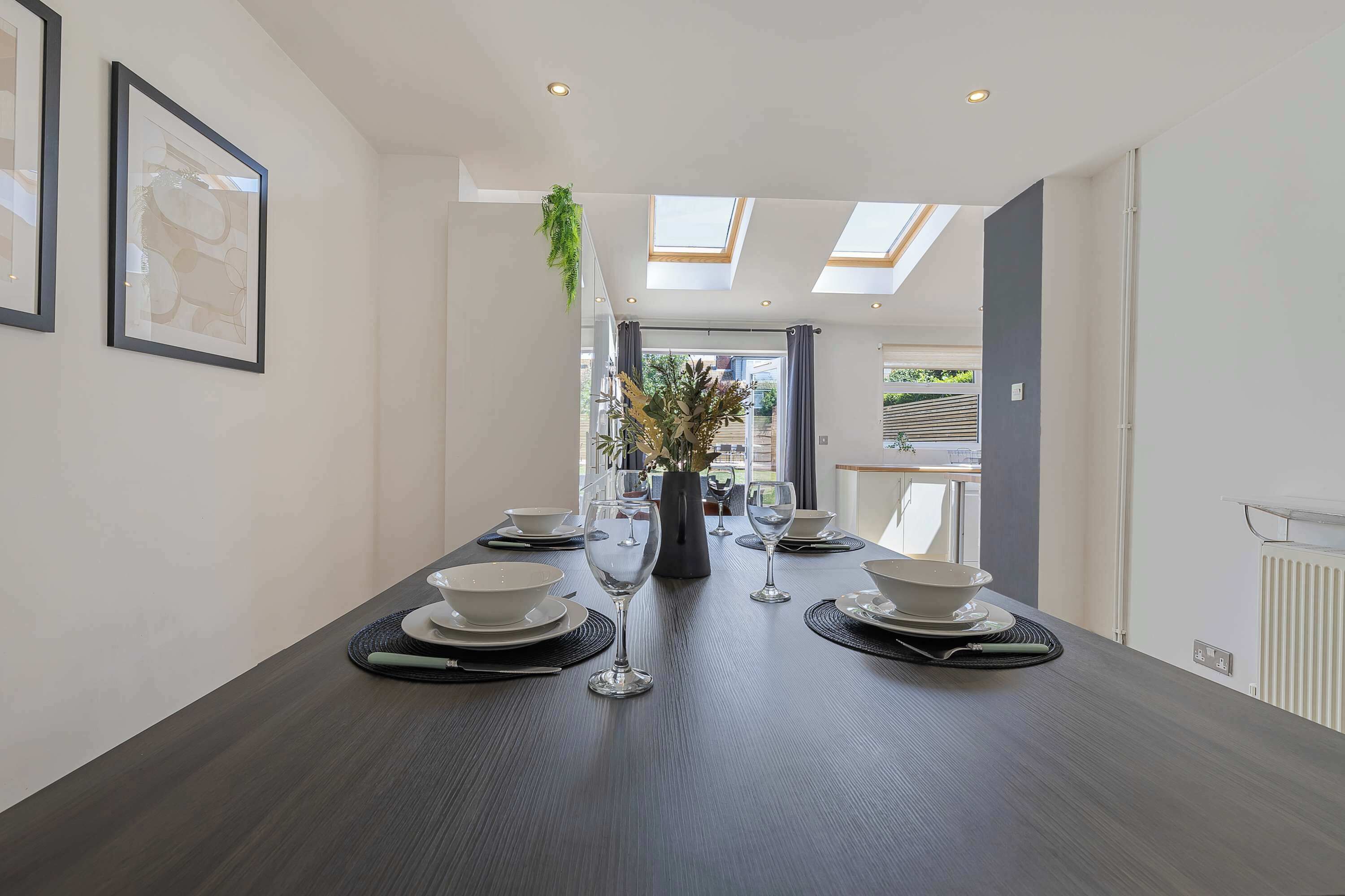 A dining room with a long table set for four, framed artwork on the walls, and a skylight above.