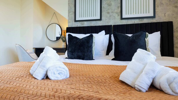 Habitación de hotel con cama, almohadas, cabecero, espejo montado en la pared y pared con dos cuadros enmarcados.