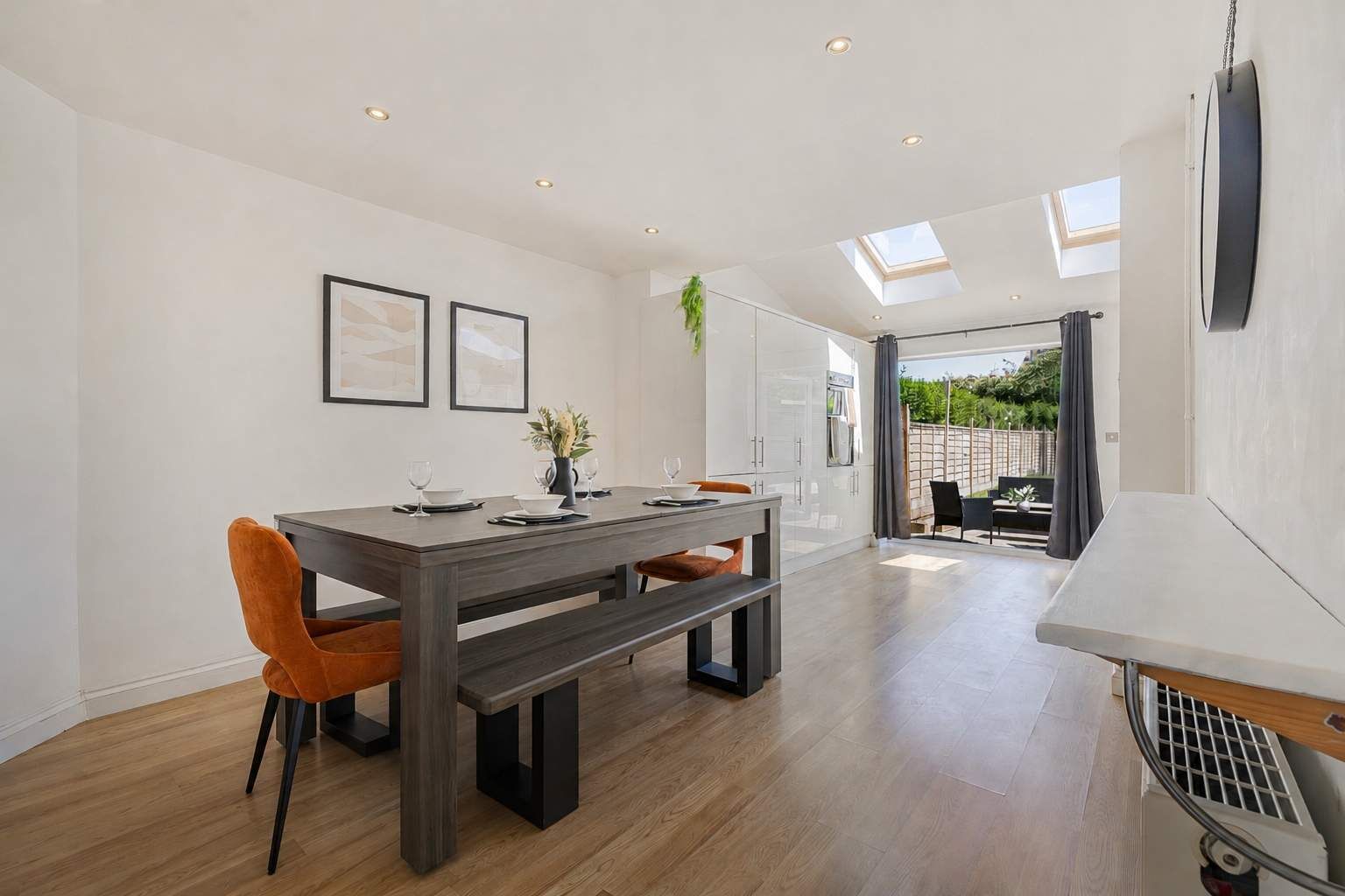 A modern dining area with a wooden table, chairs, and a bench. There are ceiling lights and a skylight.