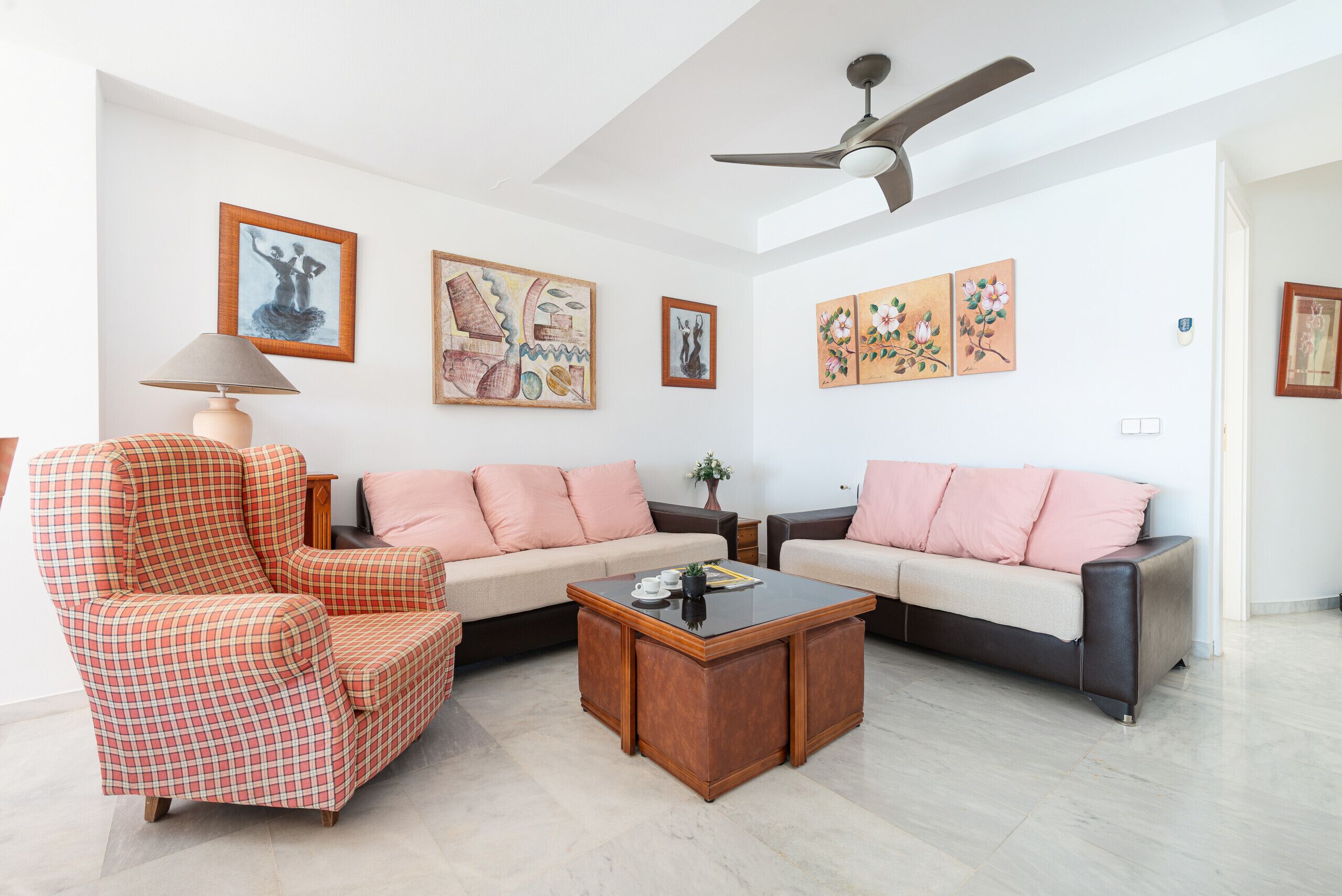A living room with a sofa, armchair, and coffee table, with artwork on the walls and a ceiling fan.