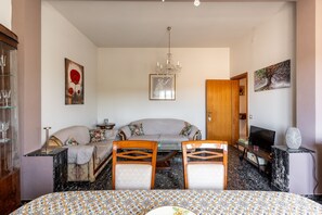 Ein Zimmer mit einer Couch, zwei Holzsesseln, einem Esstisch mit gemustertem Tischläufer, einer Kristallleuchter, einem Fernseher und Bildern an den Wänden.